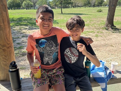 Two boys sitting on a bench in a park smiling at the camera